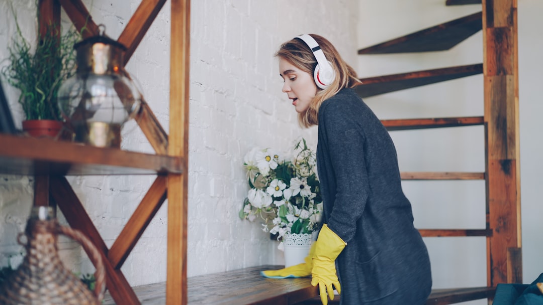 Joyful young lady is dusting the furniture, listening to music with headphones, singing and dancing having fun at home. Modern interiors, housework and happy people concept.